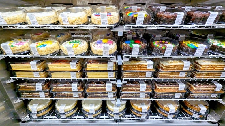 Refrigerated display shelves stocked with rows of various round cakes and rectangular dessert bars in clear plastic containers. Costco holiday hacks.