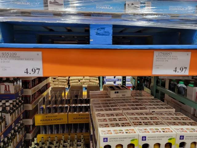 Store shelves with packaged bottles of Manuka honey and Japanese gourmet sauces, priced at 4.97 each, stacked below a blue shipping pallet. Costco pricing tricks.