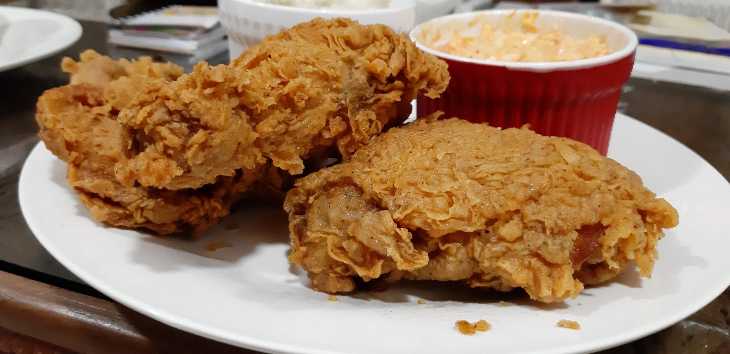 Two pieces of crispy fried chicken on a white plate next to a small red bowl of coleslaw.