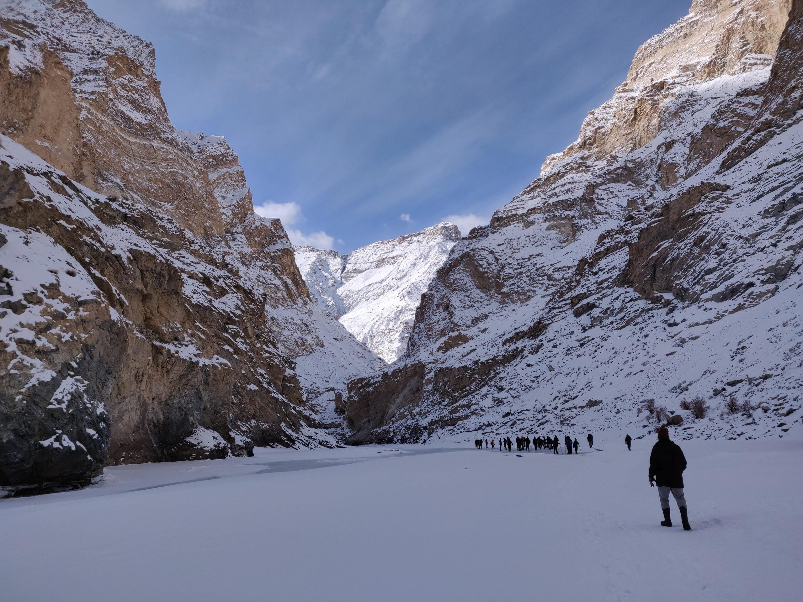 A group of people hiking across a frozen, snow-covered river flanked by high, snowy mountain walls.
