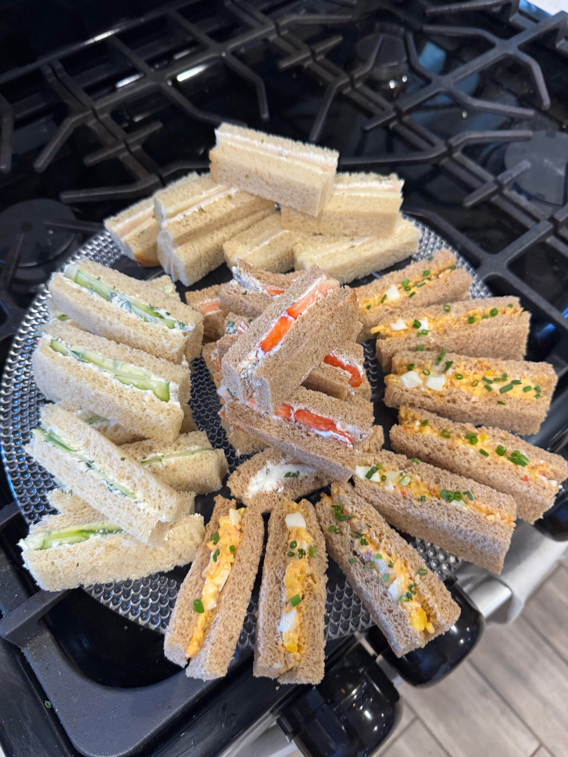 A round platter of assorted tea sandwiches, including cucumber, salmon, and egg salad, arranged on a stovetop.