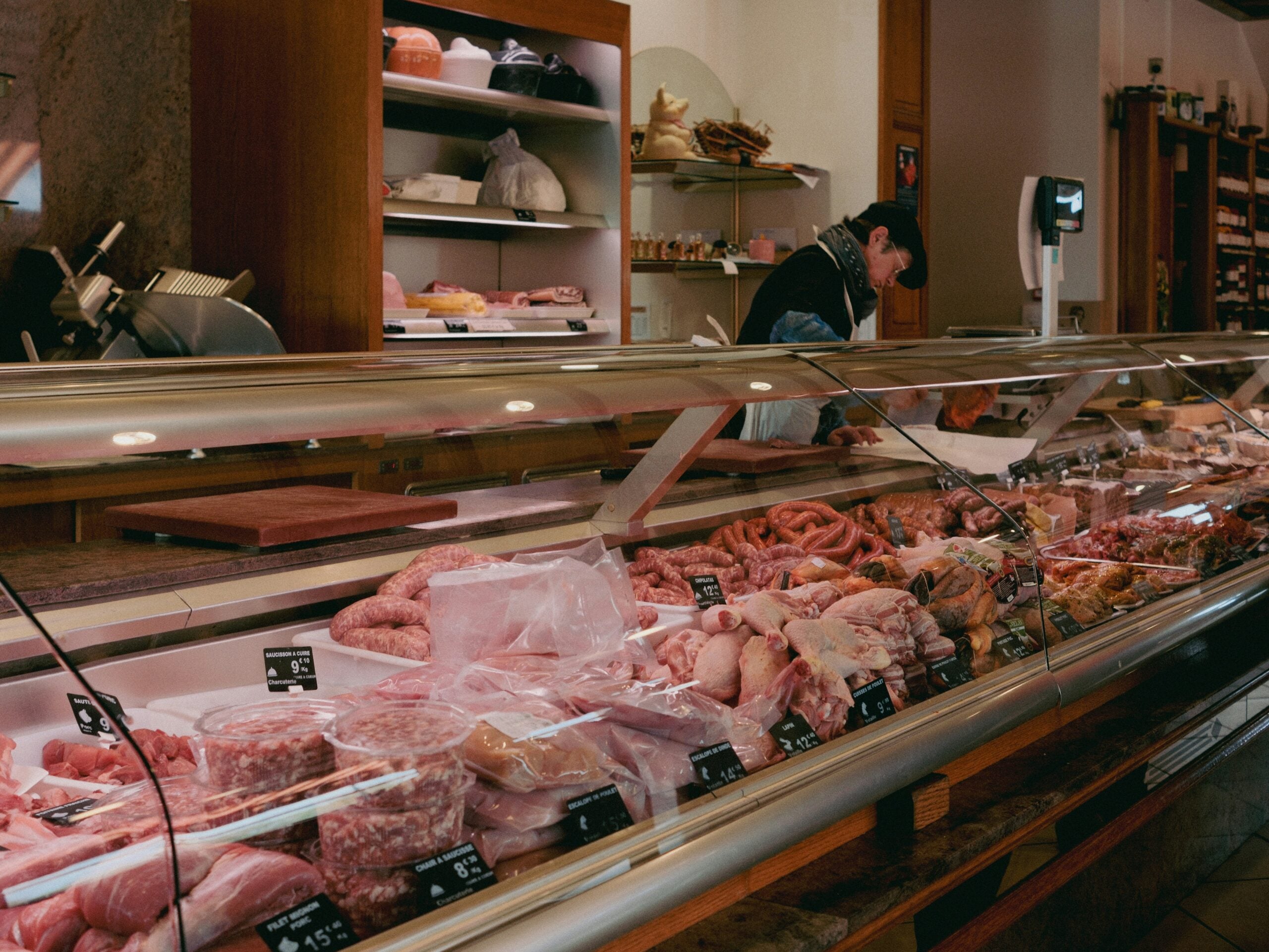 A glass display case in a butcher shop filled with various cuts of raw meat, sausages, and poultry, with a staff member working in the background.