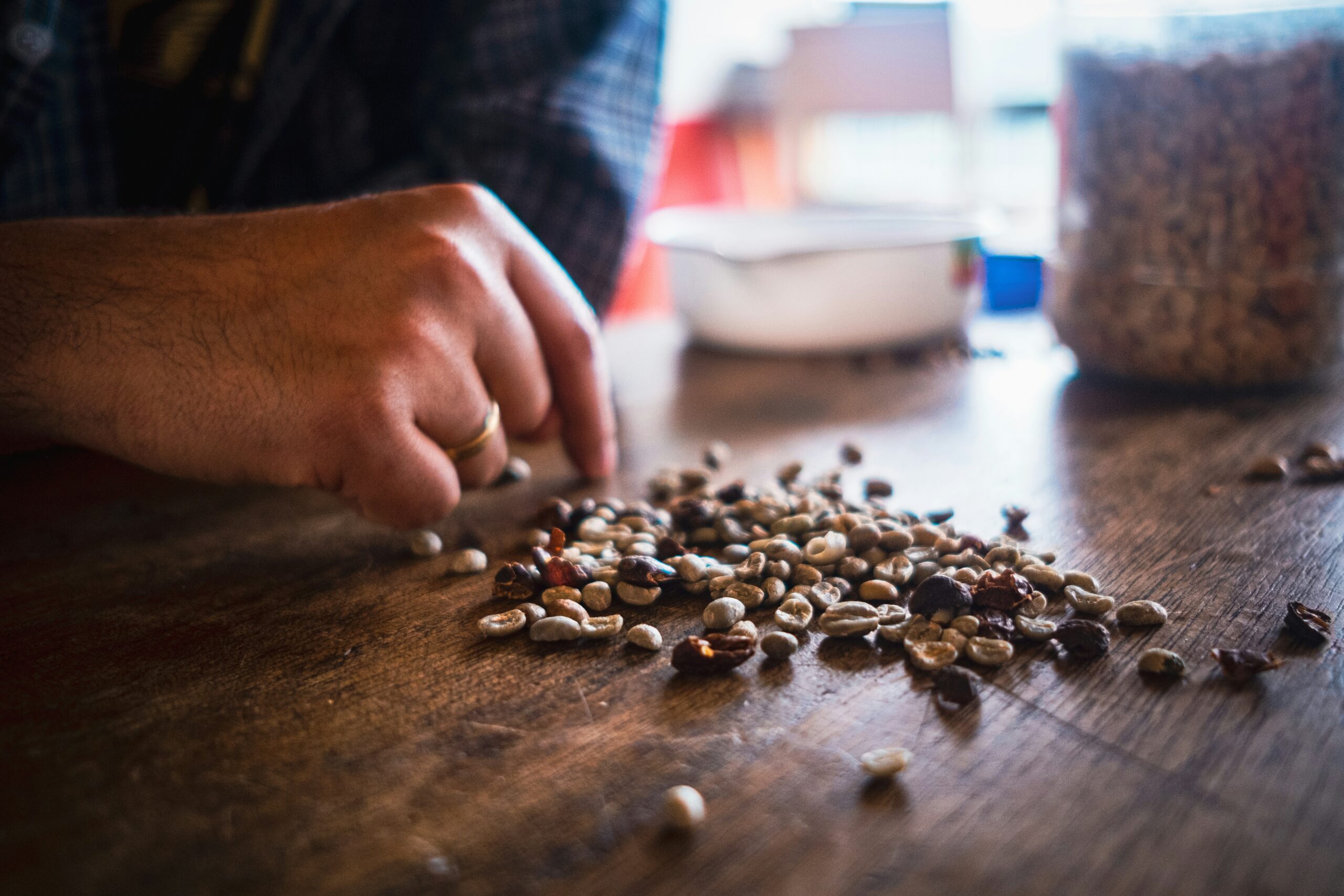 A hand with a ring sorts through a pile of mixed coffee beans on a wooden table, with a blurred container and bowl in the background.