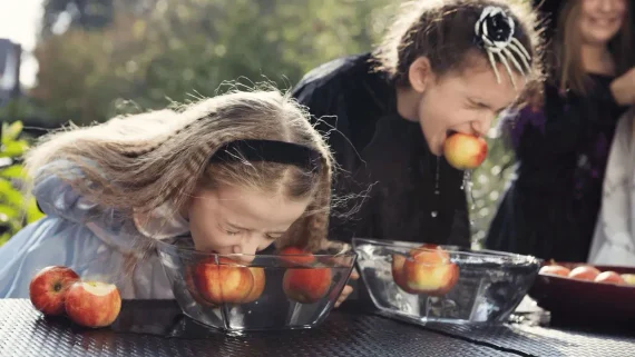 Two children in Halloween costumes bob for apples in clear bowls of water.