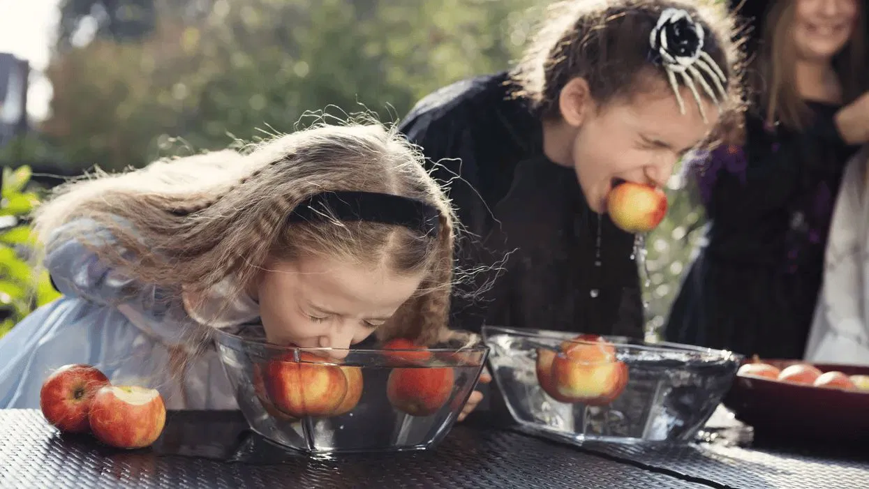 Two children in Halloween costumes bob for apples in clear bowls of water.