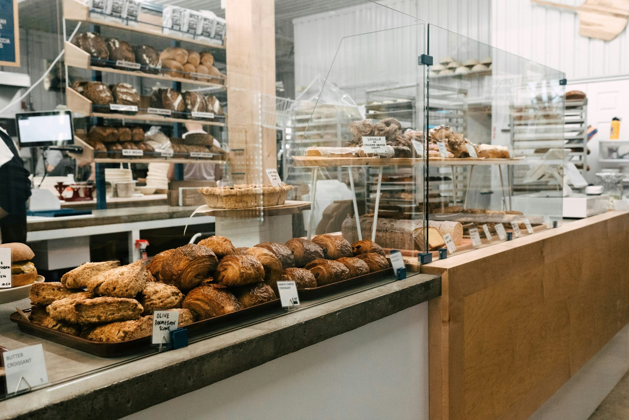Bakery counter displaying trays of croissants and scones with small price labels, with shelves of various breads visible in the background.