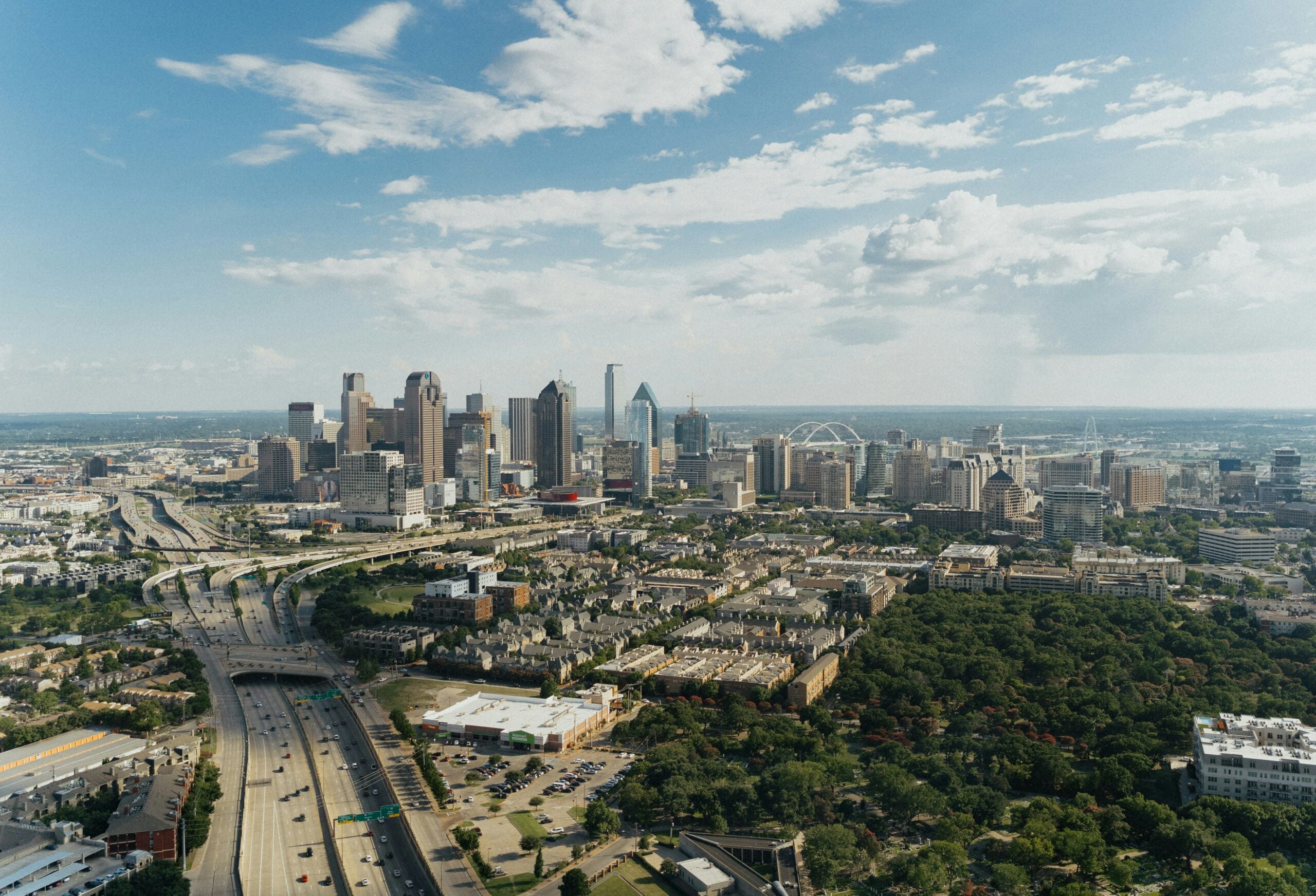 Aerial view of the Dallas skyline with a large park in the foreground and a multi-lane highway curving through the city.