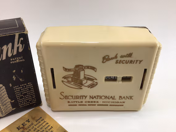 Vintage cream-colored plastic Security National Bank piggy bank with a calendar date window, next to its original box and instruction card.