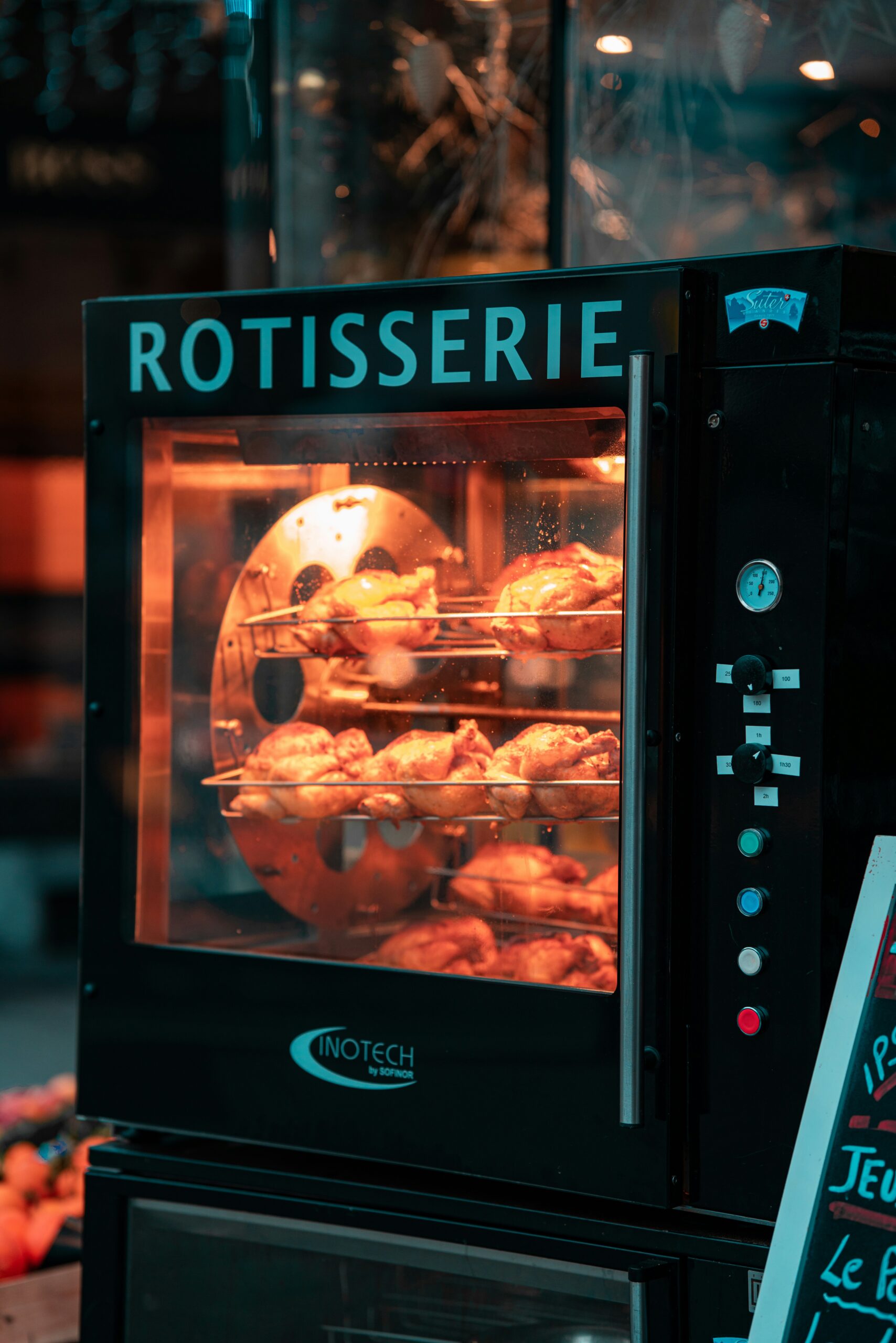 A black rotisserie oven with multiple roasted chickens inside is displayed in a restaurant or shop window. The word “ROTISSERIE” is illuminated at the top of the oven.