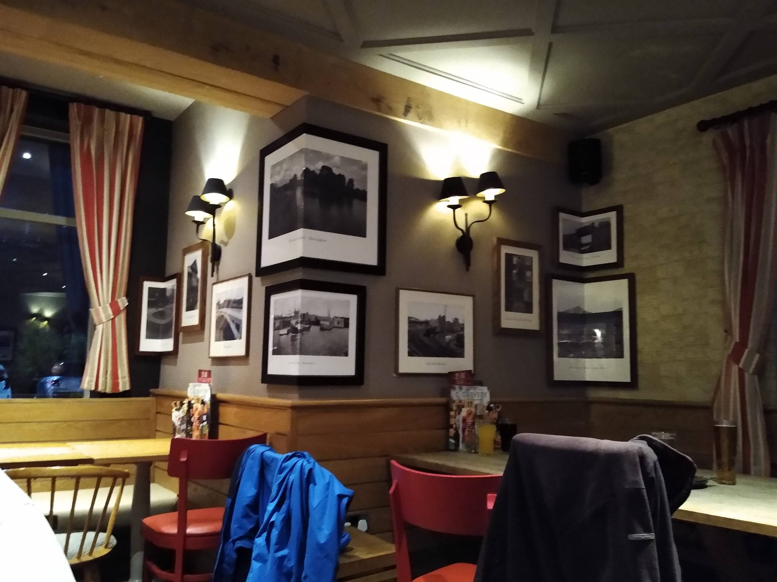 A cozy cafe corner with framed black-and-white photos on the wall, red chairs, a table, and jackets draped over chairs. Soft lighting and striped curtains add to the warm ambiance.