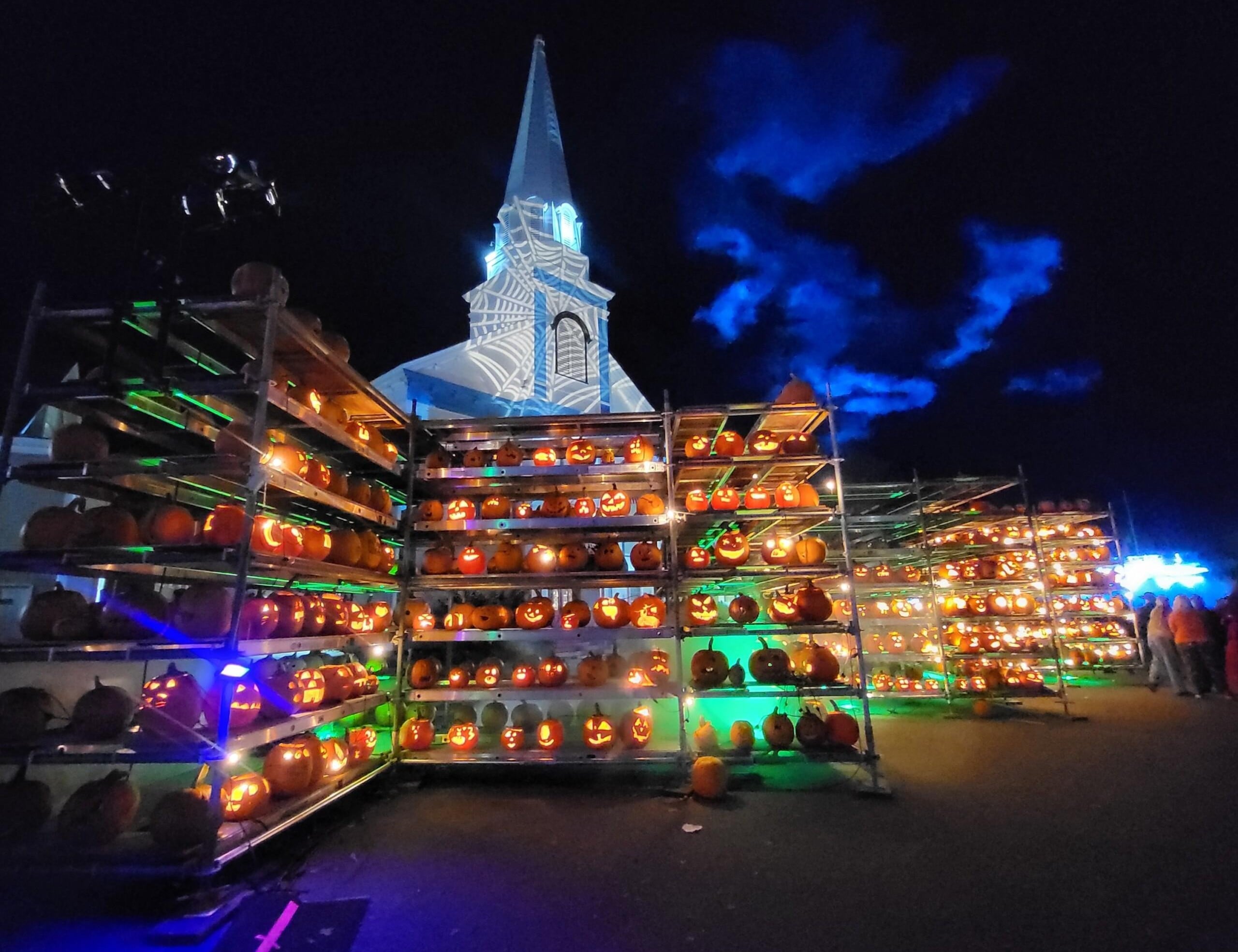 Rows of illuminated jack-o’-lanterns displayed on metal shelves in front of a church building lit with a projection of spiderwebs at night.