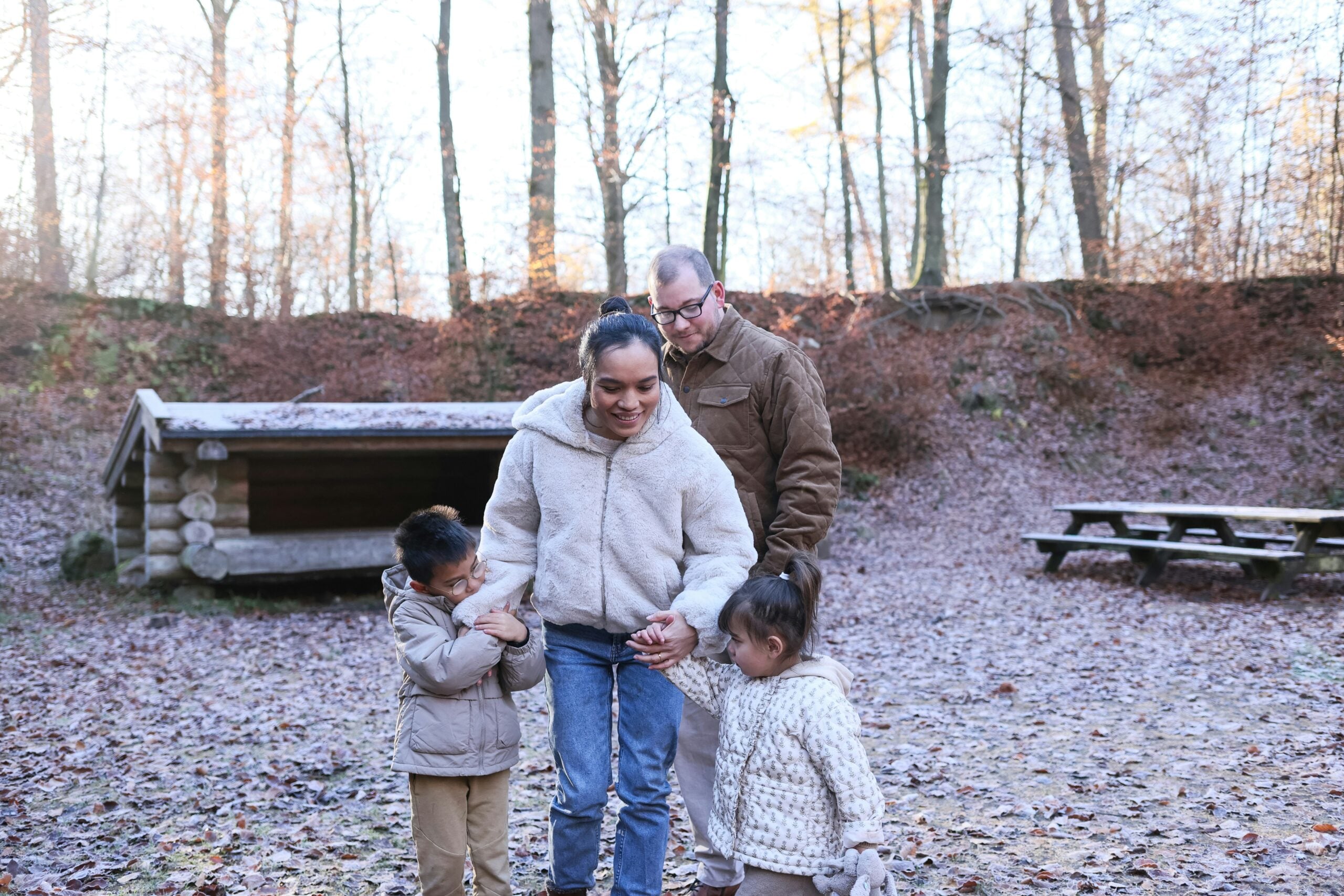 A family of four, dressed in winter clothes, walks outdoors on a frosty day. Trees, a wooden shelter, and a picnic table are in the background. The parents smile as they hold their children’s hands.