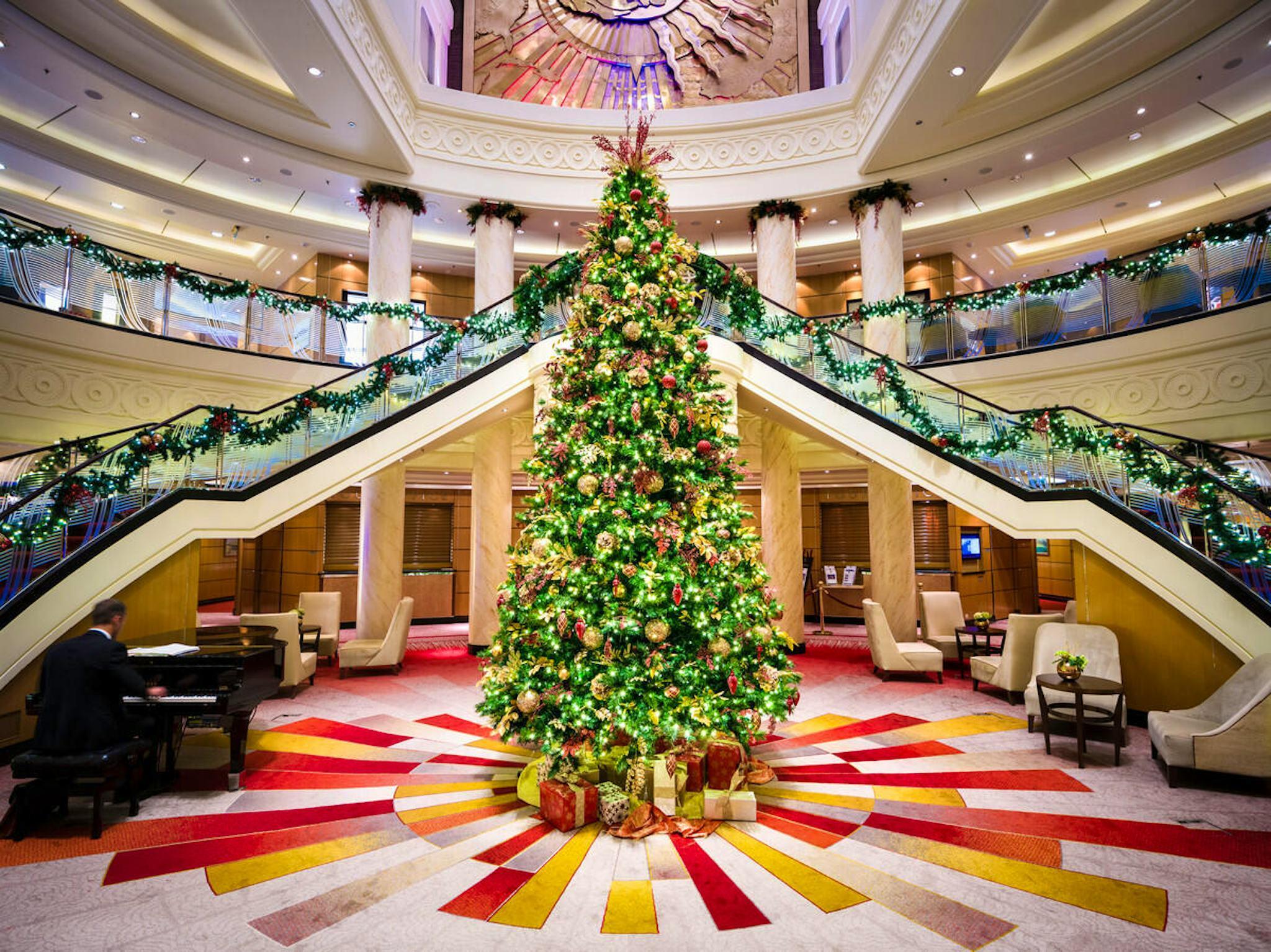 A large, decorated Christmas tree stands in the center of a grand hotel lobby, with a grand piano played by a person to the left and bifurcated staircases behind it.