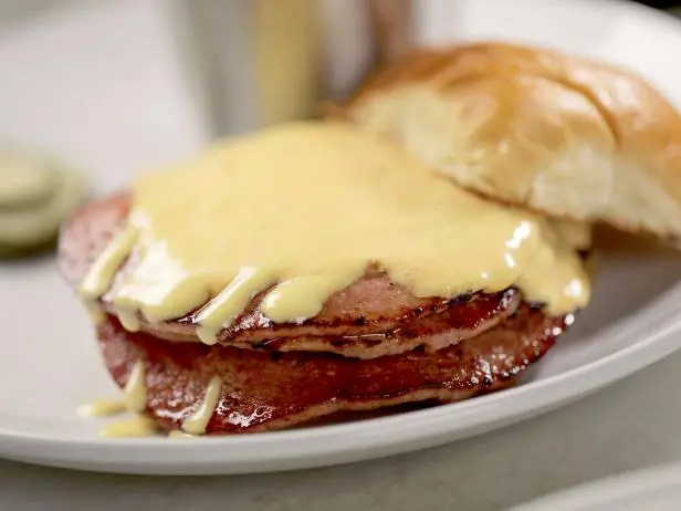 A sandwich with several slices of grilled meat topped with melted cheese sauce is served on a white plate, accompanied by a soft sandwich bun. Foods people are embarrassed to admit they love.