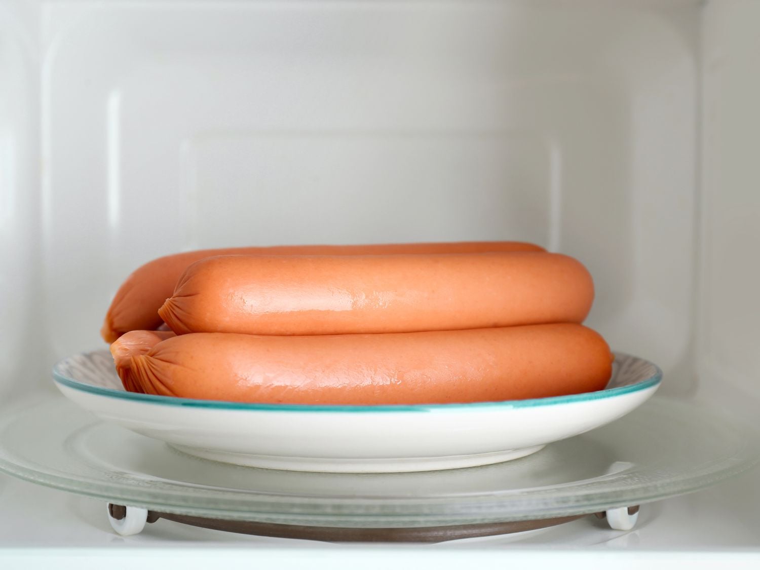 Three uncooked hot dogs on a white plate inside a microwave oven, sitting on the microwave’s glass turntable.
