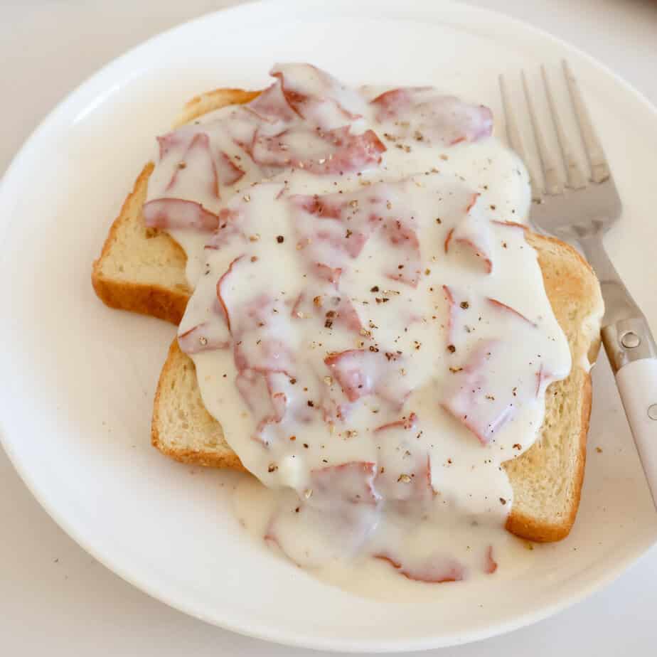 Two slices of toast topped with creamy white gravy and pieces of chipped beef, sprinkled with black pepper, on a white plate with a fork.