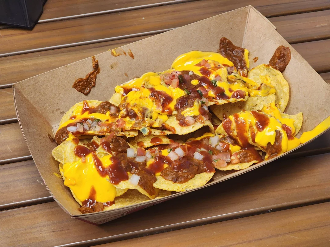 A cardboard tray filled with tortilla chips topped with melted cheese, chili, diced onions, tomatoes, corn, and a drizzle of sauce, sitting on a wooden table.