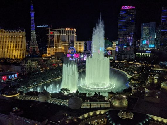Aerial night view of the illuminated Bellagio Fountains in Las Vegas, with the Eiffel Tower replica and surrounding hotel casinos glowing in the background.