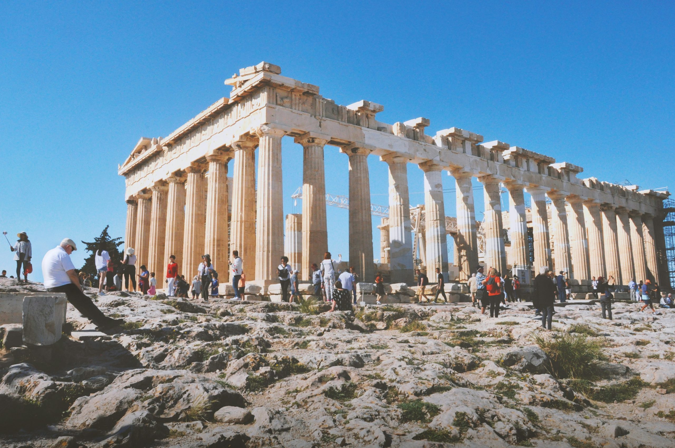 Tourists explore the Parthenon in Athens, walking along the rocky ground in front of the ancient marble columns.