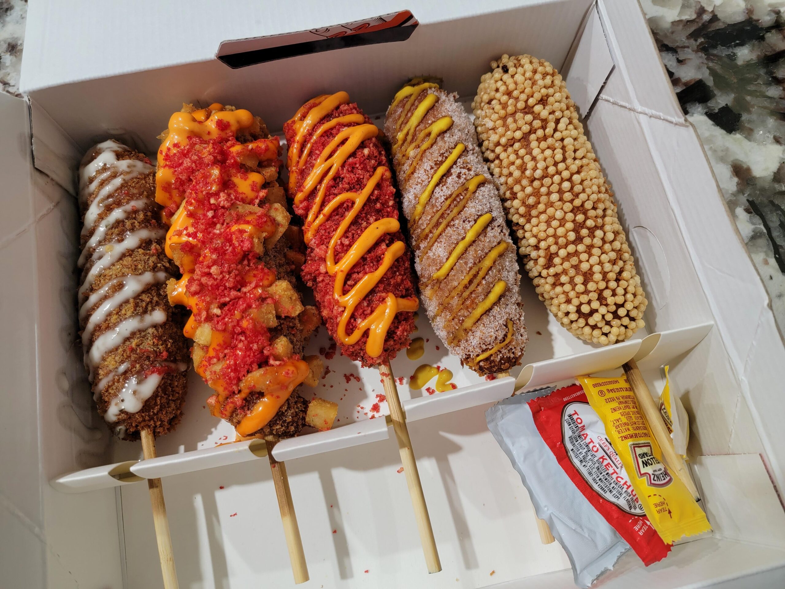Five Korean corn dogs with various colorful toppings are arranged in a white box, along with ketchup and mustard packets on the side. The corn dogs have different coatings, including sauces, crumbs, and cheese.