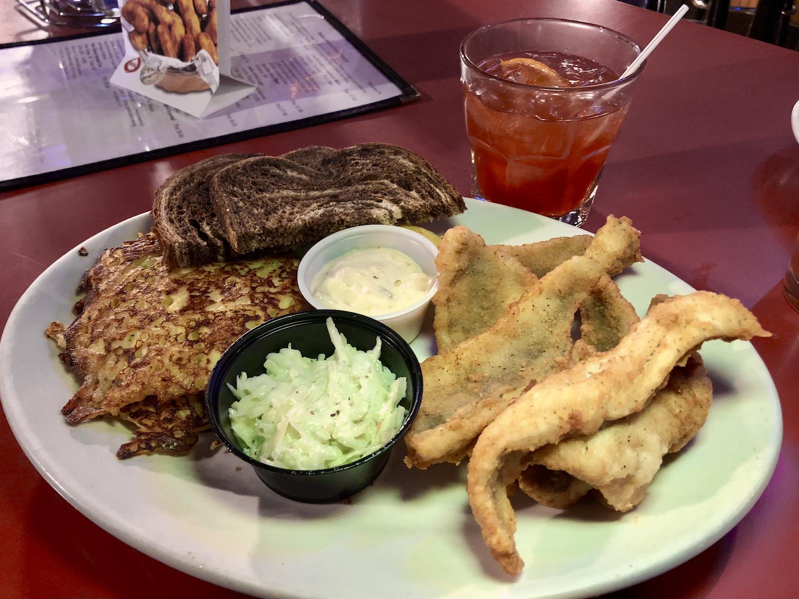 A plate of fried fish, a potato pancake, two slices of rye bread, coleslaw, and tartar sauce, served with an iced cocktail.