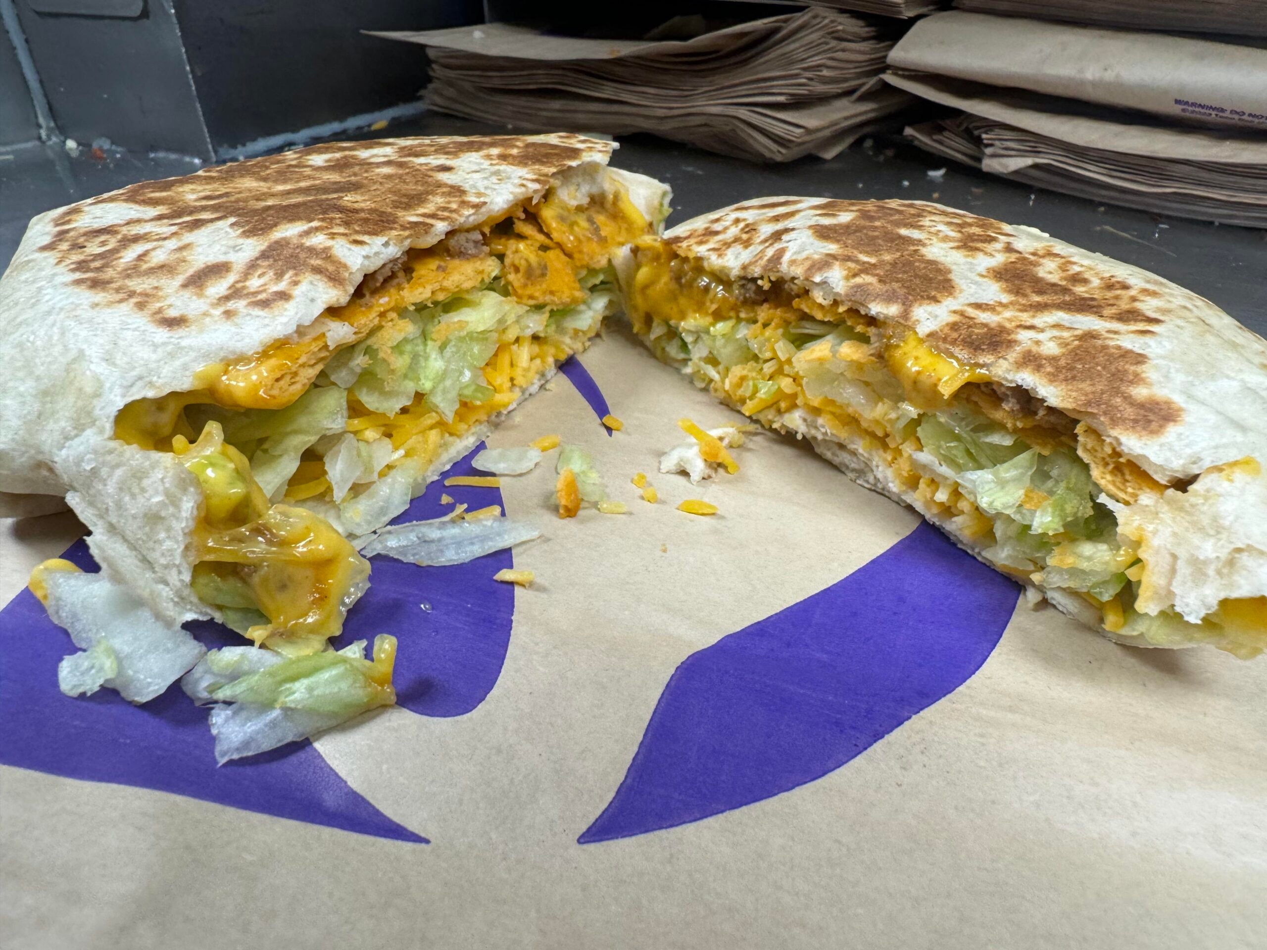 A grilled flour tortilla crunchwrap cut in half, revealing layers of meat, melted cheese, shredded lettuce, and a crispy tostada shell.