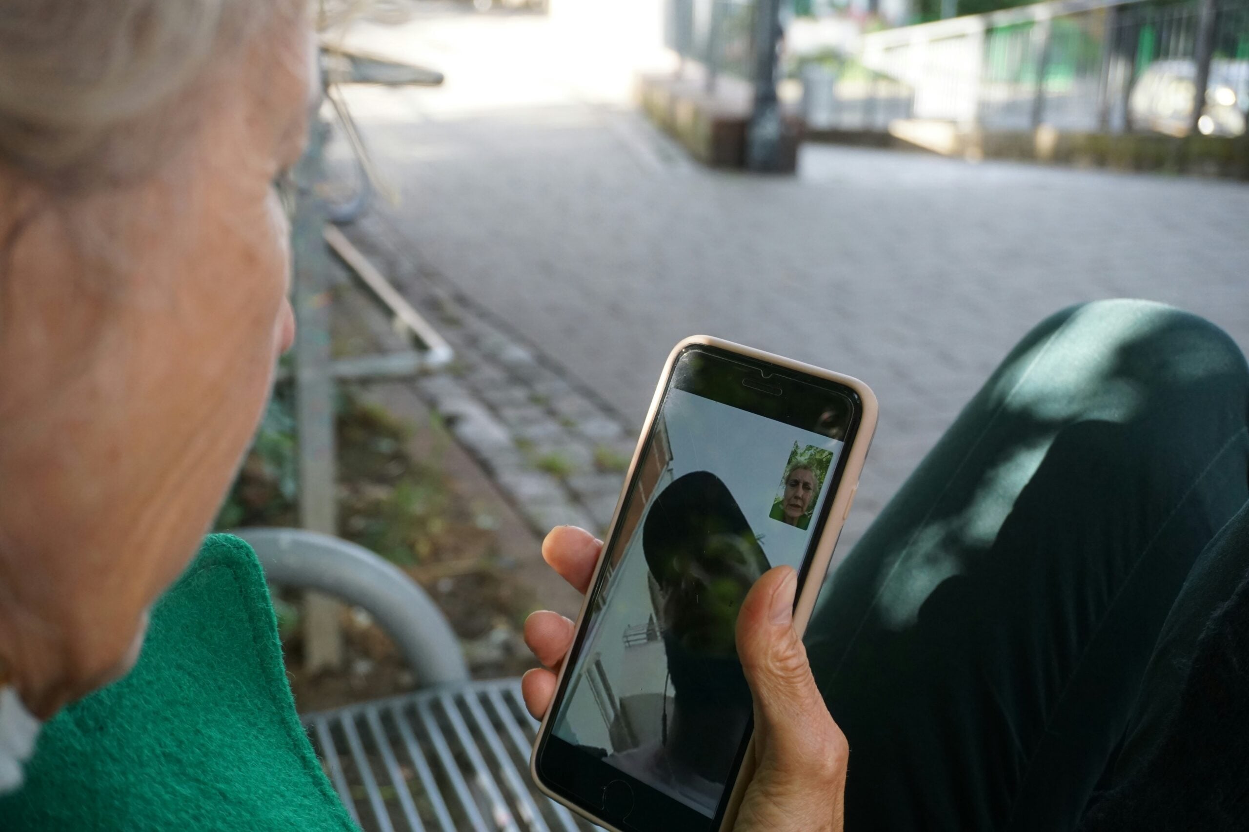 An older adult sits on a bench outdoors, holding a smartphone and video calling someone. The screen shows the face of the person they are talking to.