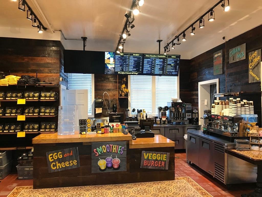 The interior of a cafe with a wooden counter featuring signs for egg and cheese, smoothies, and veggie burgers, with a digital menu board overhead and shelves of retail products on the left.