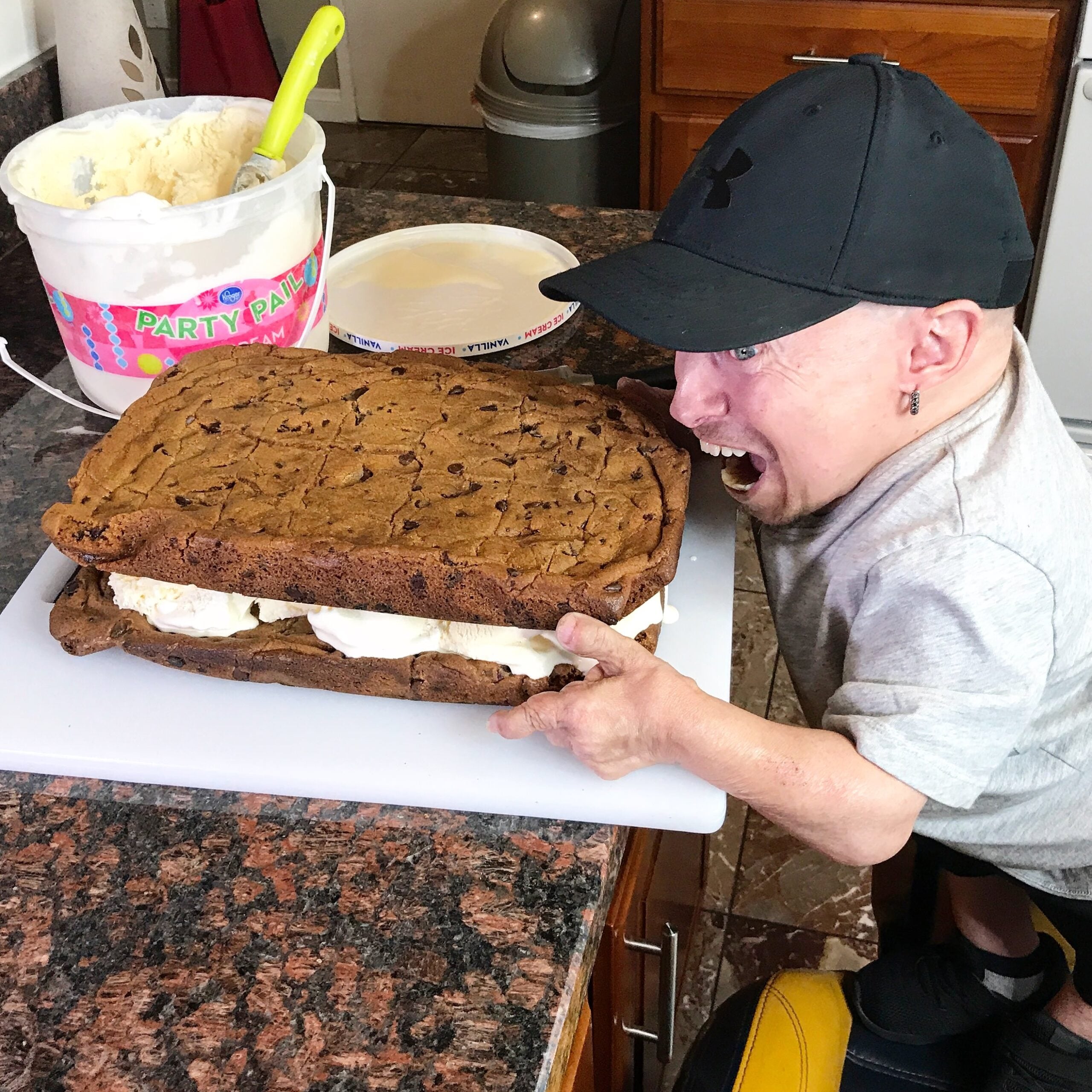 A person in a black cap leans in to take a bite of a giant ice cream sandwich made with two large chocolate chip cookies.
