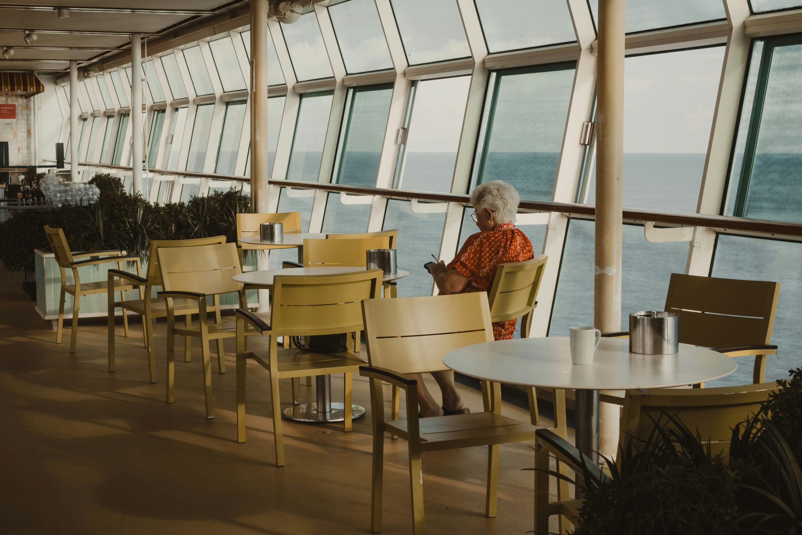 An elderly person sits alone at a yellow table near large windows on a ship, looking at their phone with empty chairs around them and the ocean visible outside.