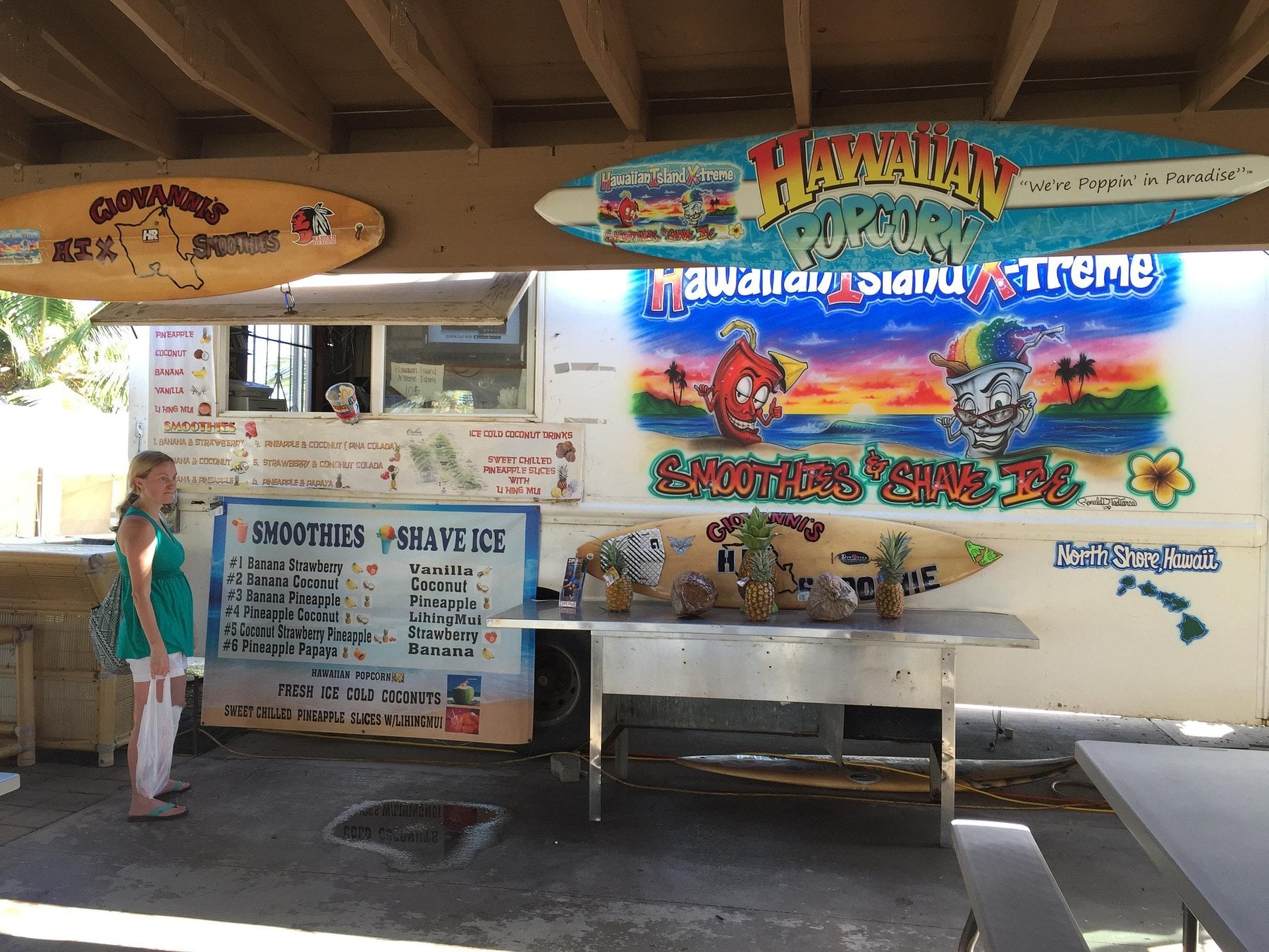 A woman stands at a Hawaiian food truck with menus for smoothies, shave ice, and popcorn, featuring surfboards as signage.