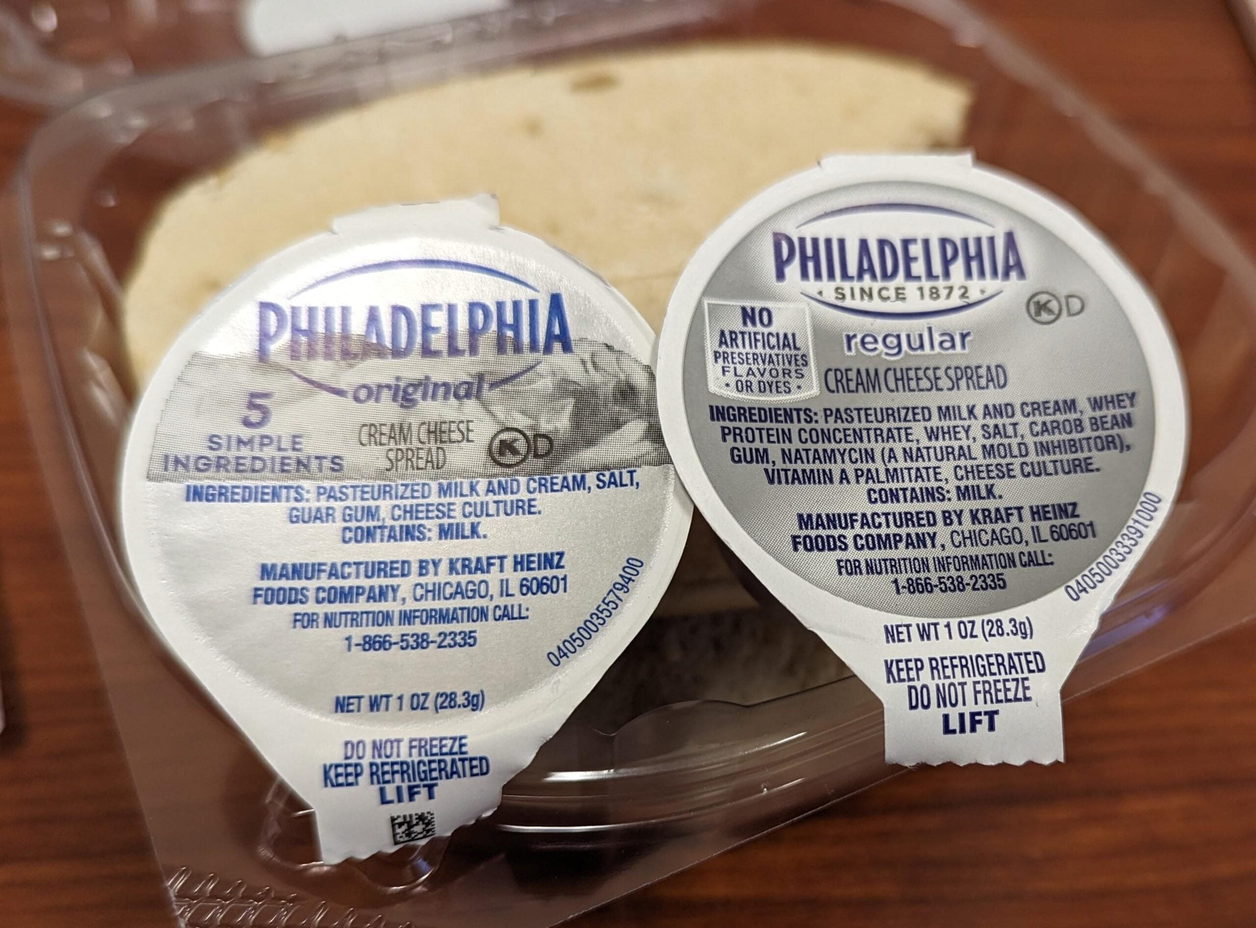 Two individual 1 oz. Philadelphia cream cheese tubs, one labeled Original and one Regular, sitting on top of a plastic container.