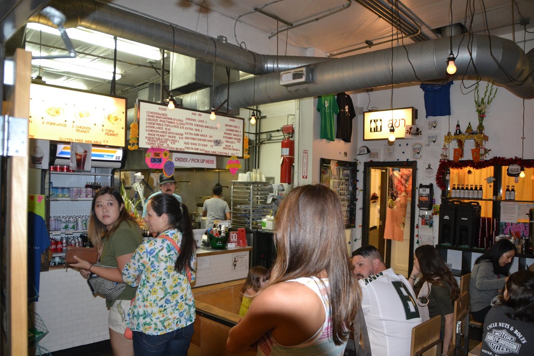 Customers wait at the order counter of a restaurant with a printed menu board displayed above the kitchen.