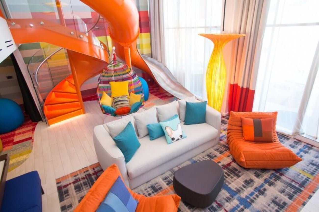 A living room with a white sofa, orange lounge chair, hanging egg chair, and an orange slide connecting to a spiral staircase, highlighting unique cruise ship amenities.