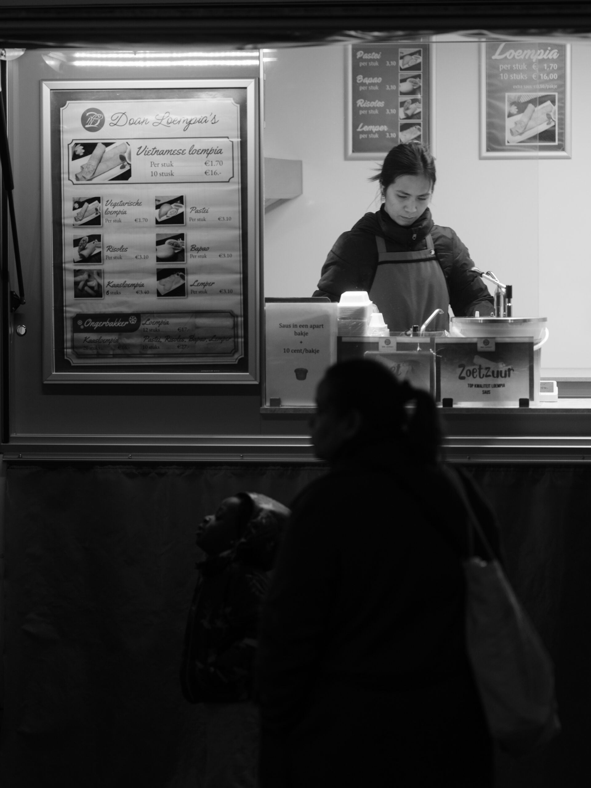 A woman works at a food stall preparing dishes, while another person stands in the foreground. The scene is in black and white.