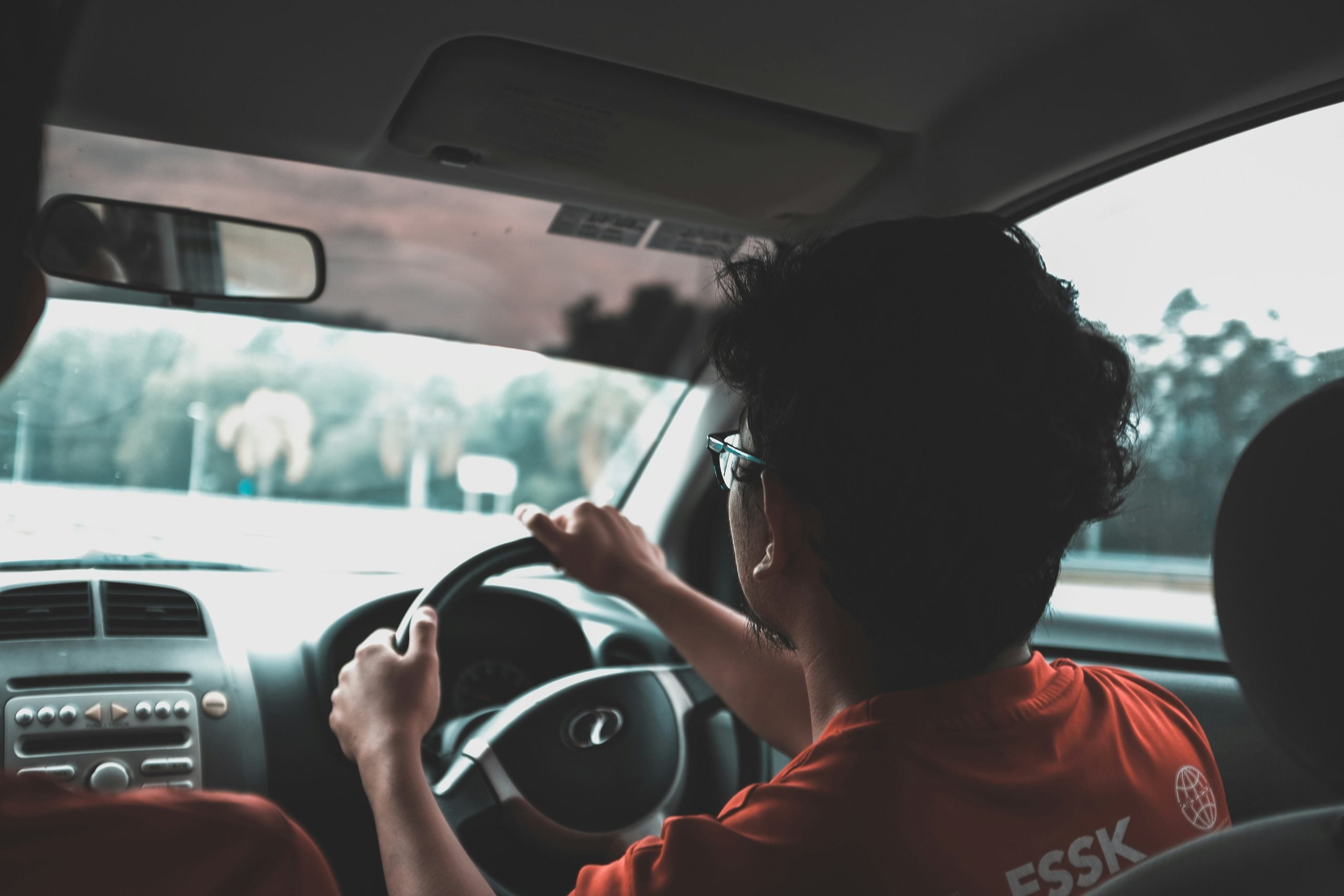 A person wearing glasses and an orange shirt driving a car.