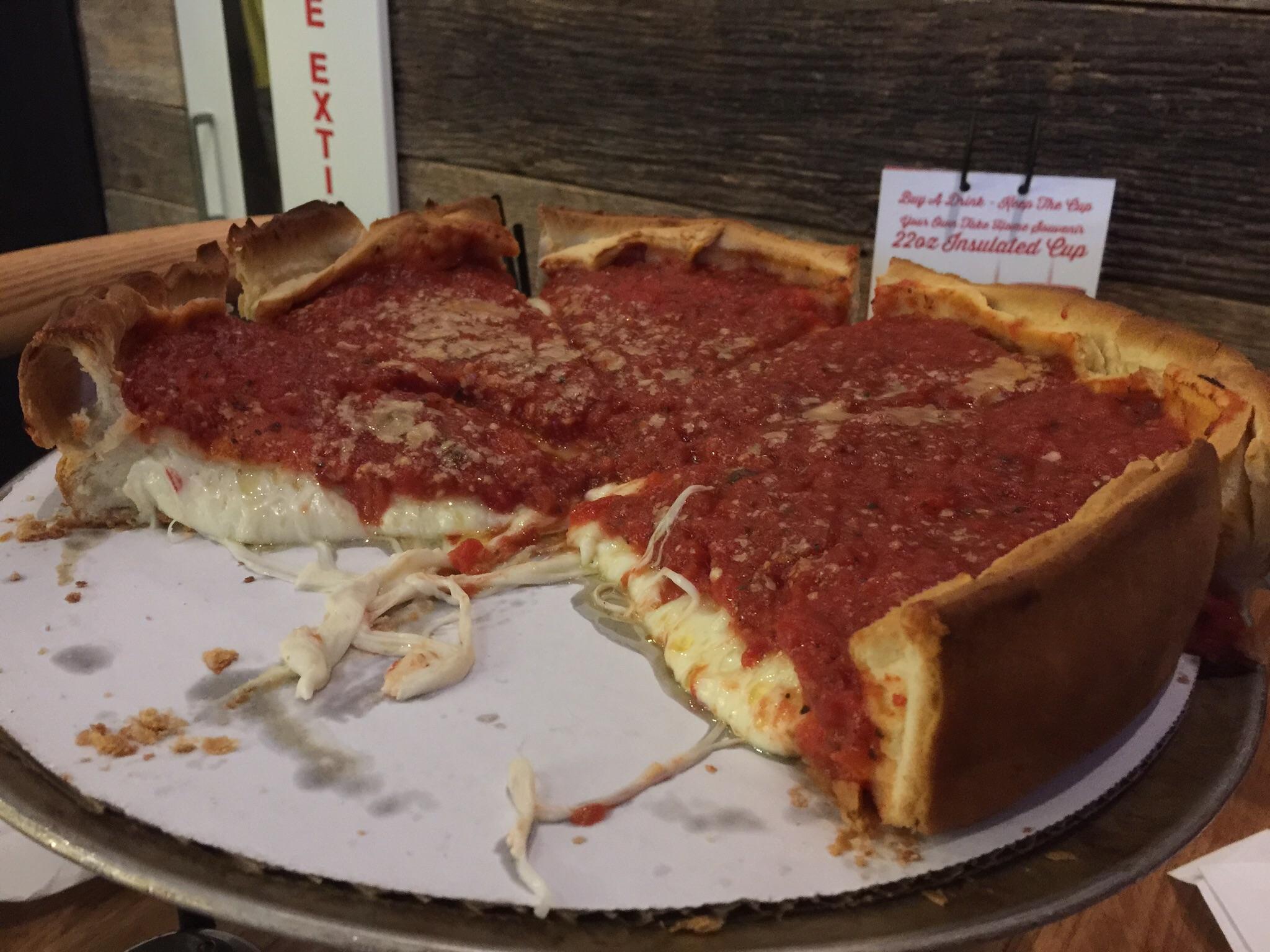A partially eaten Chicago-style deep-dish pizza sits on a metal tray, revealing thick layers of cheese and tomato sauce inside a golden, high-edged crust.