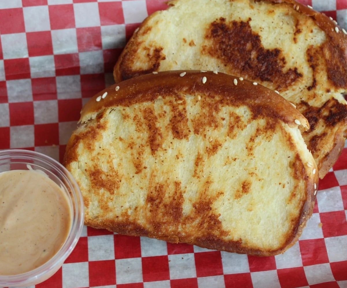 Two slices of toasted bread sprinkled with sesame seeds, served with a small cup of dipping sauce on red and white checkered paper.