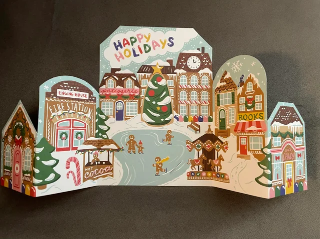 A tri-fold holiday card illustration featuring a winter village scene with a fire station, a Christmas tree in a town square, gingerbread figures, a carousel, and a candy shop.