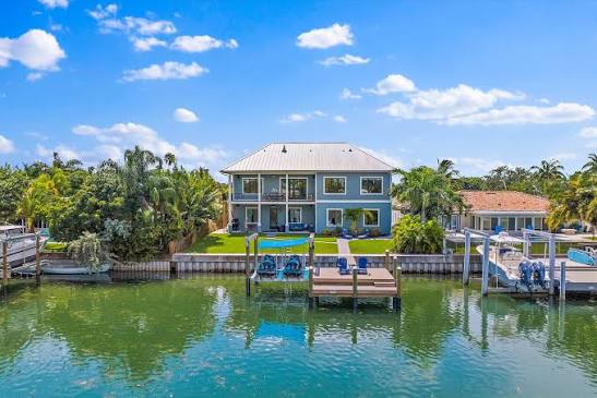 A large two-story house with a dock, lawn, and palm trees, sits beside a calm waterfront under a bright blue sky with scattered clouds.