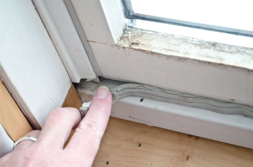 A hand points to cracked, old caulking on a window frame, showing signs of wear and needing repair. The frame appears dirty and weathered.