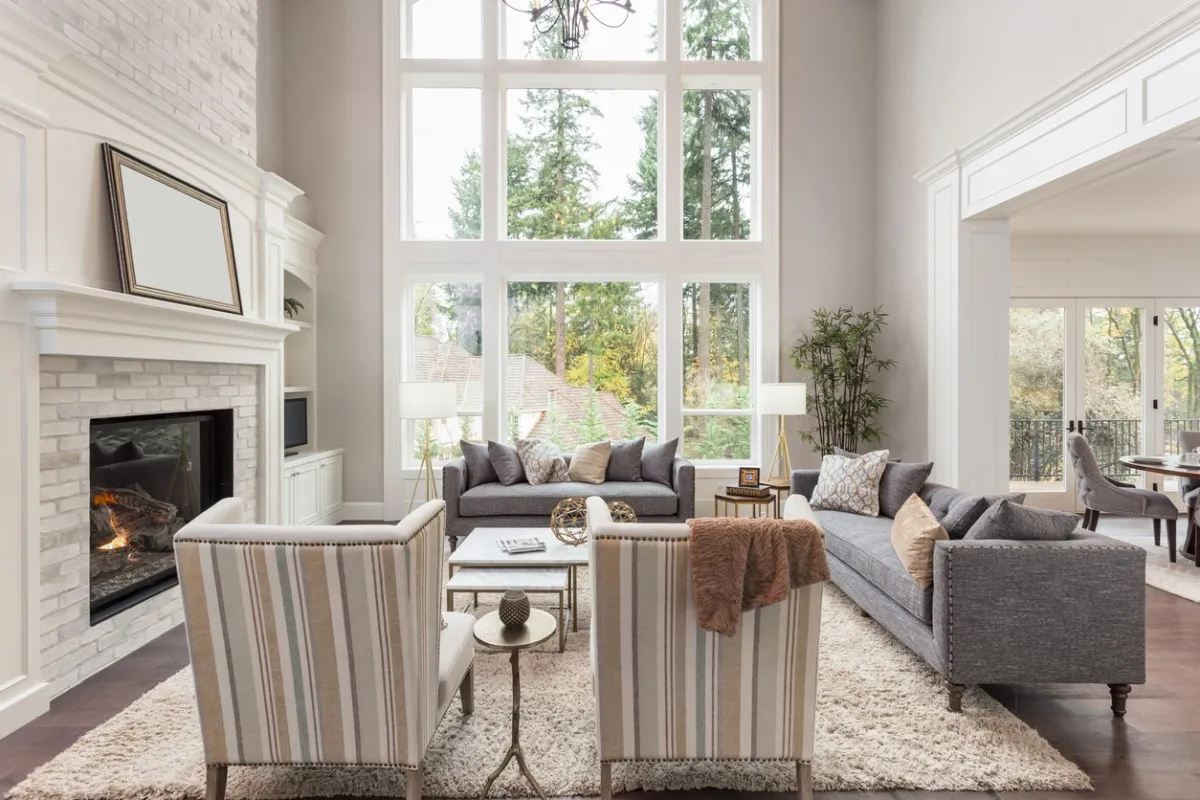 Bright, modern living room with tall windows, gray sofas, striped armchairs, a fireplace, and a view of trees outside. Neutral decor and cozy accents create an inviting atmosphere.