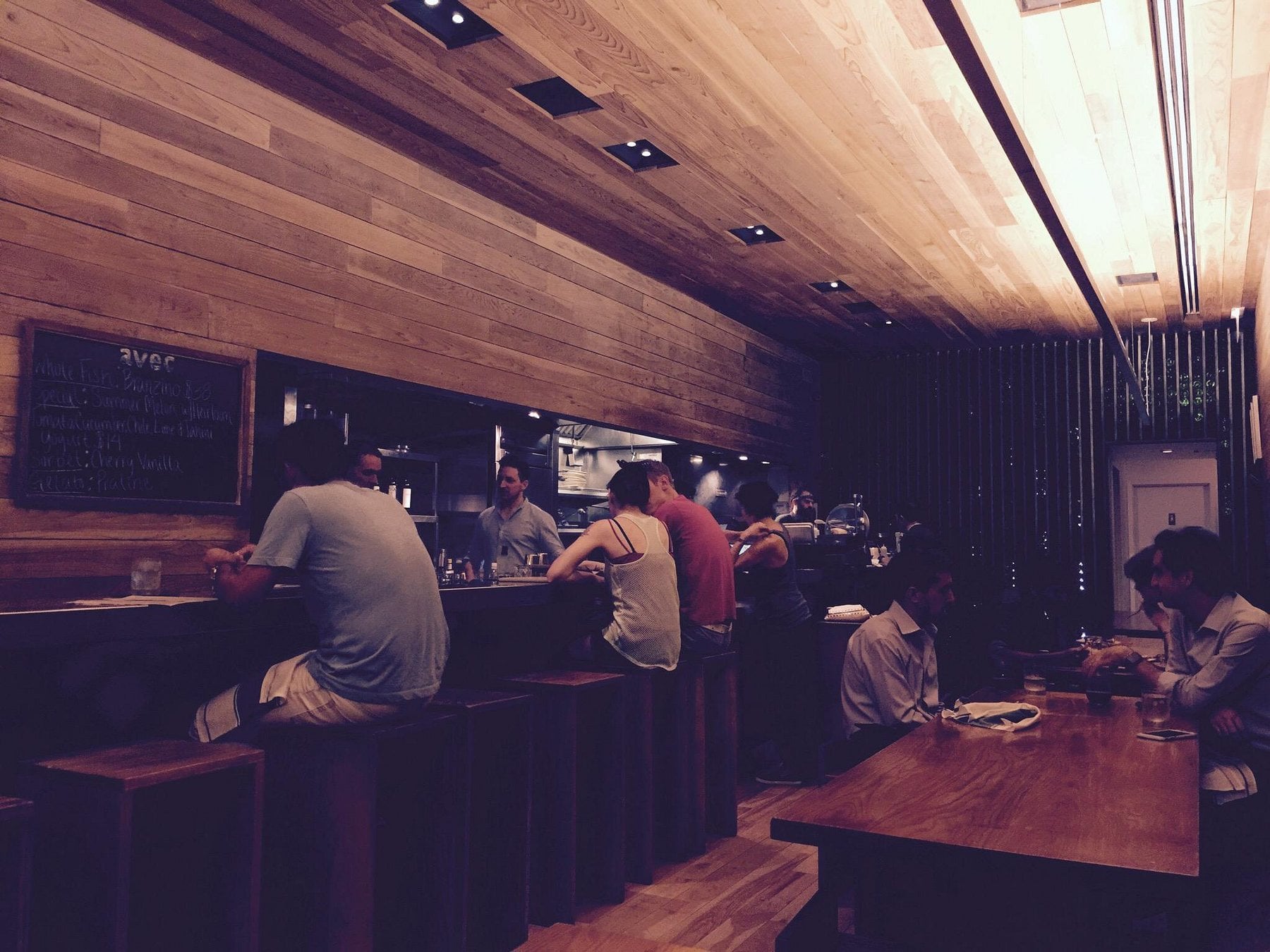 People sitting at bar seating and dining tables in a restaurant with wood-paneled walls and a chalkboard menu.