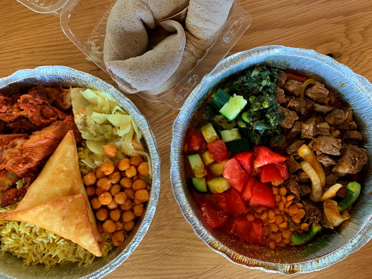 Two foil takeout containers of Ethiopian food and a side of injera bread on a wooden table.
