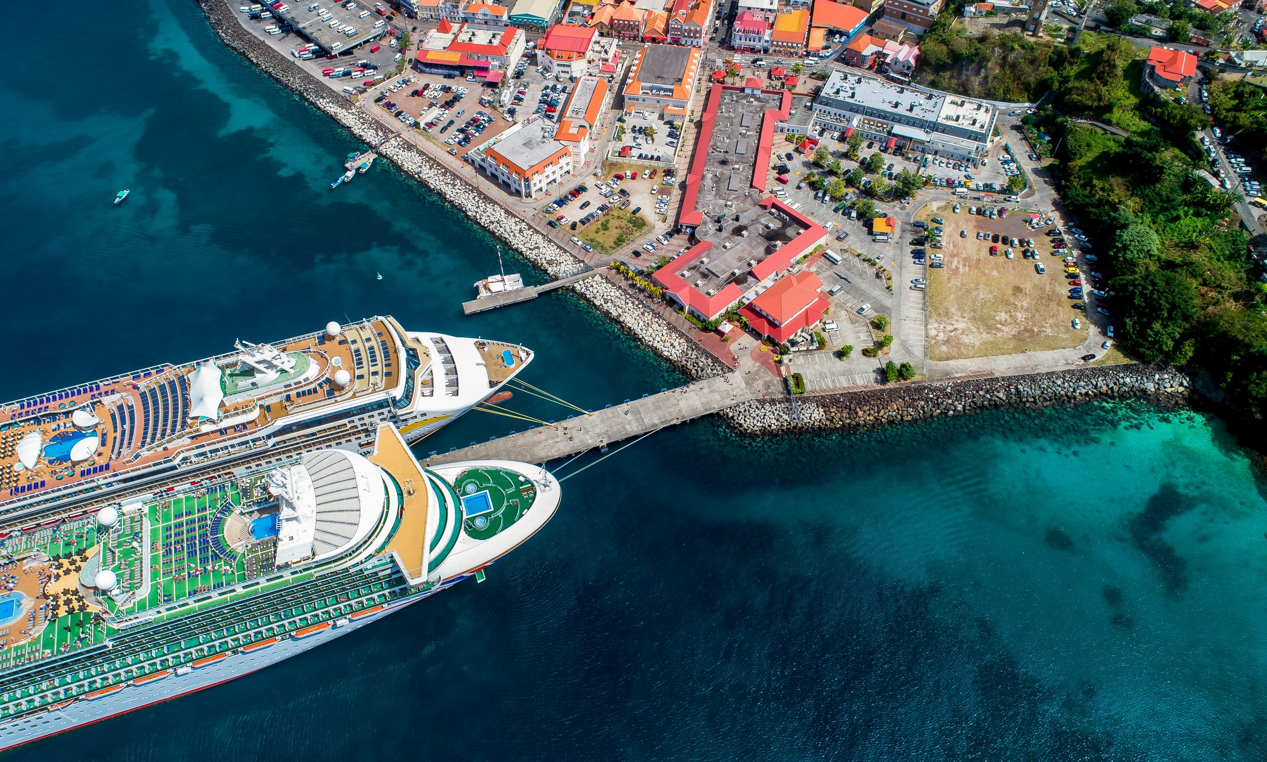 Aerial view of two large cruise ships docked at a pier next to a coastal town with colorful buildings and clear blue-green water.