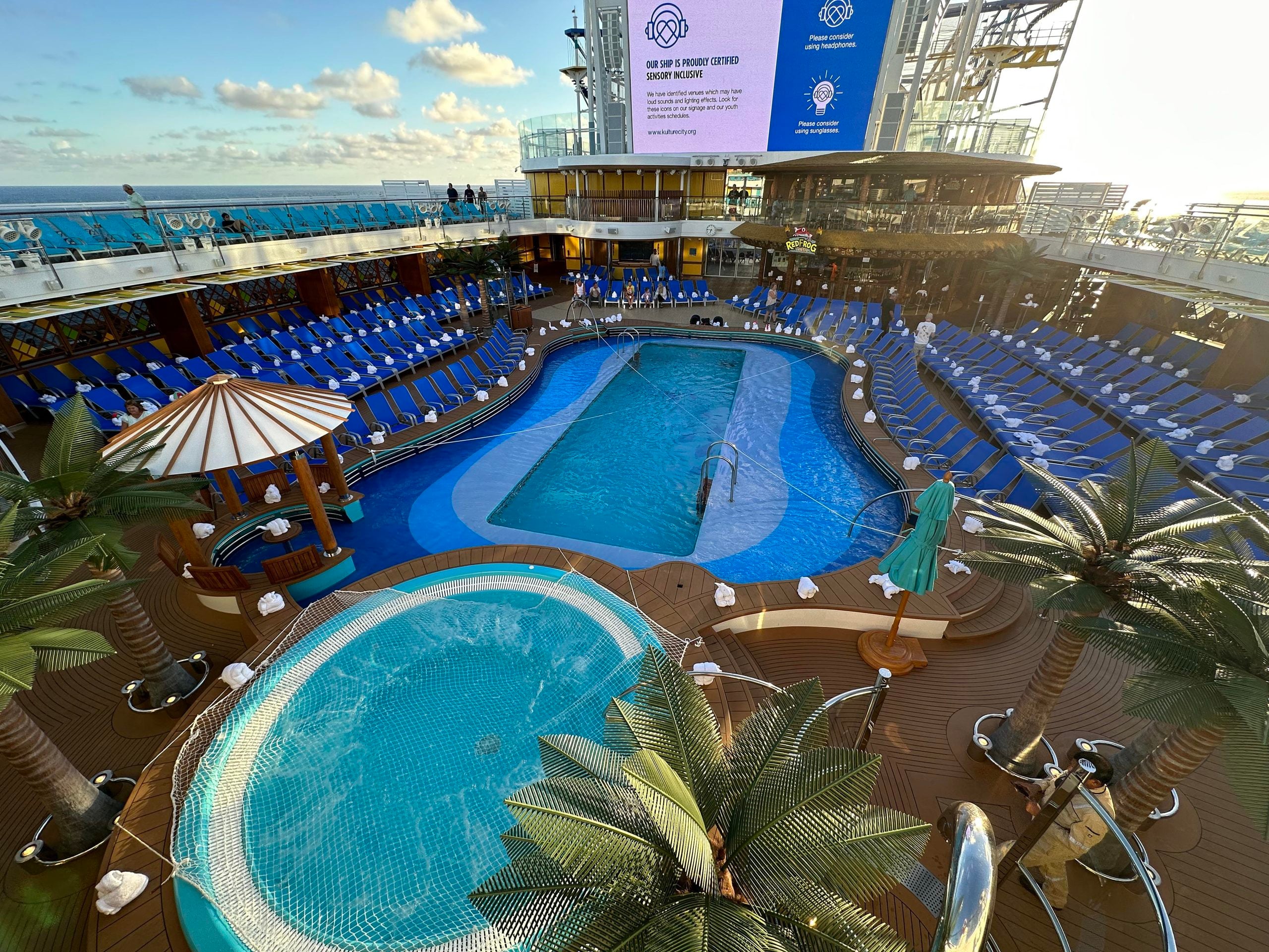 An elevated view of a cruise ship pool deck featuring two swimming pools, rows of blue lounge chairs, and a large sign announcing that the ship is sensory inclusive.