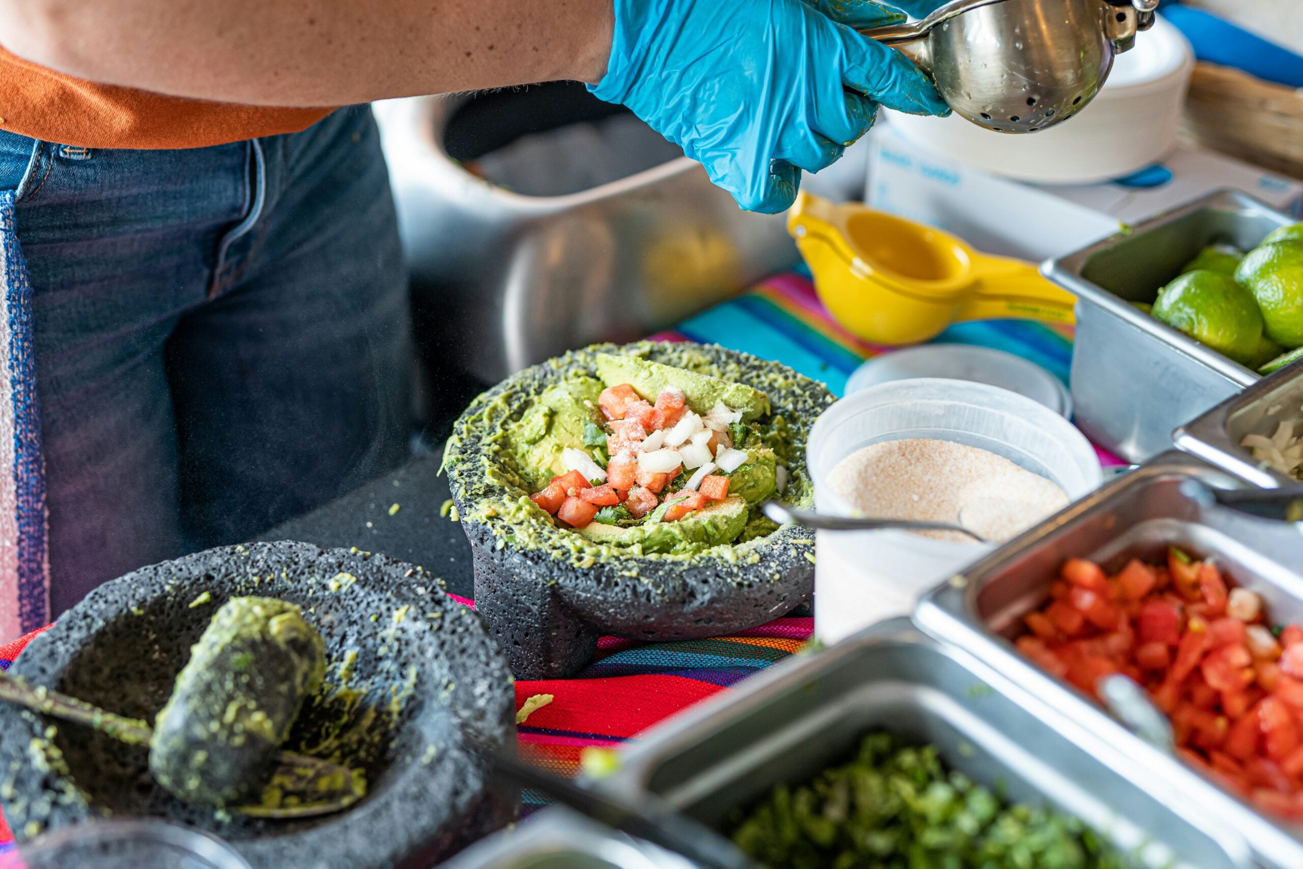 A person wearing blue gloves squeezes a lime over a stone bowl of guacamole topped with diced tomatoes and onions.