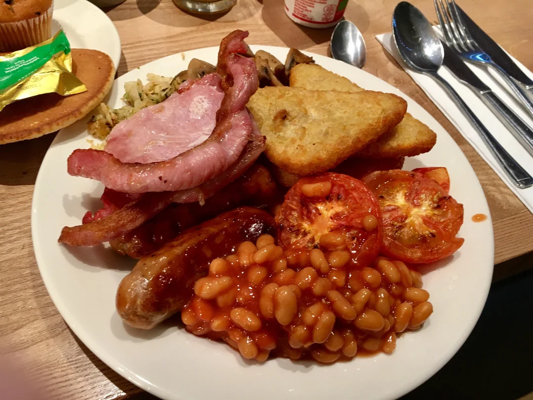 A full English breakfast on a white plate featuring a sausage, bacon, baked beans, grilled tomato halves, and hash browns.