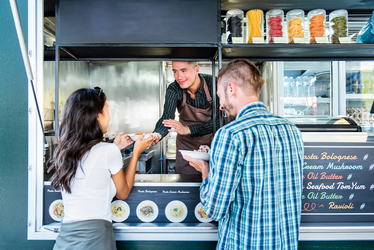 A food truck worker in an apron hands a bowl of pasta through the takeout window to a customer as another waits, with a menu featuring pasta dishes displayed on the counter.