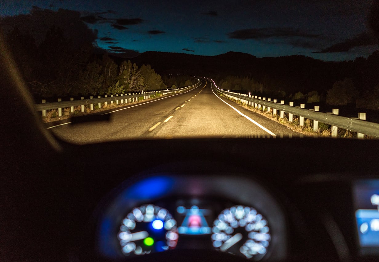 A view from inside a car at night, looking down a dark, winding road illuminated by headlights.